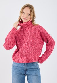 Maglione rosso lavorato a maglia con collo alto a imbuto, polsini e orlo a coste. Presenta un motivo testurizzato e una vestibilità ampia. Abbinato a jeans azzurri chiari.