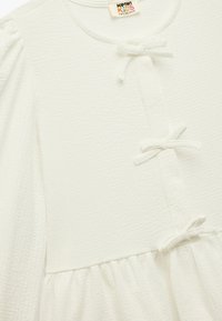 Robe blanche pour enfants en tissu texturé, avec un col rond et des manches bouffantes douces. Présente un devant à boutons et des nœuds décoratifs.