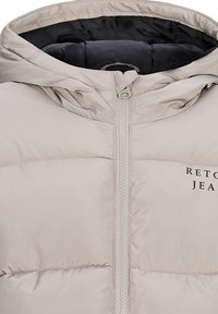 Jasno beżowa kurtka puchowa z kapturem, z zamkiem błyskawicznym z przodu i przeszyciami. Czarna podszewka wewnętrzna. Logo "RETOUR JEAN" z boku.