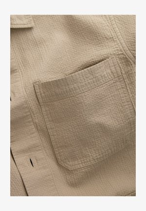 Beige textiel met een gestructureerd patroon, met een vierkante zak aan de linkerkant, zichtbaar stiksel en een subtiel ruitpatroon.