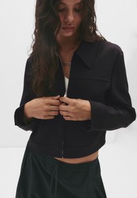 Jachetă neagră scurtă cu guler și închidere cu fermoar, combinată cu pantaloni negri largi. Materialul pare neted, cu un design structurat.