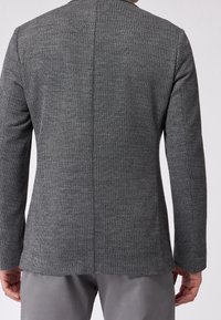 Uomo che indossa un blazer grigio a coste, sagomato, abbinato a pantaloni grigi chiari, fotografato di spalle su uno sfondo neutro.