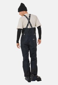 Zwarte snowboard overall met verstelbare banden, voorzien van logo-accenten, ritszakken en contrasterende stoffenpanelen. Gedragen over een beige shirt.