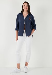 Navy button-up shirt with rolled sleeves, featuring a small embroidered accent, paired with white jeans and white sneakers.
