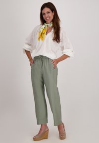 Witte oogjesblouse met een ontspannen pasvorm, gecombineerd met lichtgroene broek met koordjes, en een gele geprinte sjaal. Sleehaksandalen maken de outfit compleet.