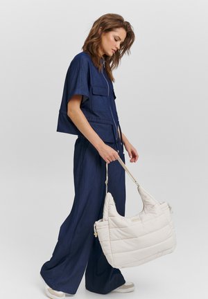 Marineblaues, kurzes Oberteil und weite Hosenbeine aus weichem Denimstoff; das Model hält eine cremefarbene, gesteppte Handtasche mit strukturierter Form und Schulterriemen.