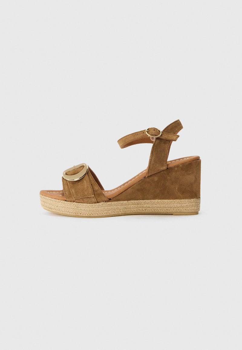 Sandali con zeppa in suede marrone, con punta chiusa, cinturino alla caviglia e accentuazione circolare dorata. La base avvolta in juta aggiunge texture e altezza.