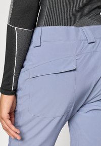 Pantaloni de culoare albastru deschis, cu o textură netedă, dotati cu un buzunar la spate și o croială ajustată. Asortați cu un top negru, texturat, cu mânecă lungă.