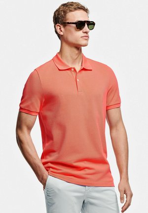 Man met een koraalrode poloshirt, witte broek en schildpaddenrandzonnebril, met zijn handen in zijn zakken, kijkt opzij tegen een witte achtergrond.