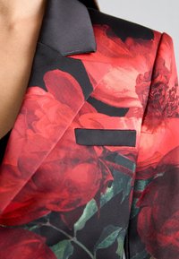 Blumenblazer mit roten Rosen auf schwarzem Hintergrund, mit einem kontrastierenden schwarzen Satin-Kragen und einer kleinen Tasche auf der Brust.