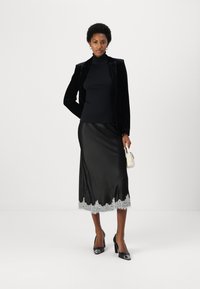 Black velvet blazer over a black turtleneck, paired with a knee-length black satin skirt featuring a lace hem. Black pointed heels and a white handbag.
