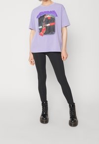 T-shirt en coton violet avec un graphisme d'un joueur de basketball et le texte "JORDAN" en caractères gras ; associé à des leggings noirs et des bottes à lacets.