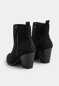 Botas de tobillo de ante negro con tacones de bloco y cremalleras laterales, vistas desde la parte posterior sobre un fondo blanco.
