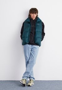 Puffer-vesti tummansininen, mustan hupparin päällä. Vaaleansiniset farkut ja beige urheilusandaalit mustilla yksityiskohdilla täydentävät asukokonaisuuden.