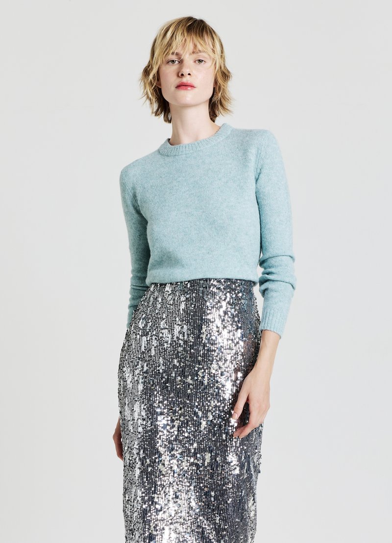 Hellblauer Wollpullover mit Rundhalsausschnitt und langen Ärmeln, kombiniert mit einem schimmernden, silbernen, vollbesetzten Rock, der das Licht reflektiert.