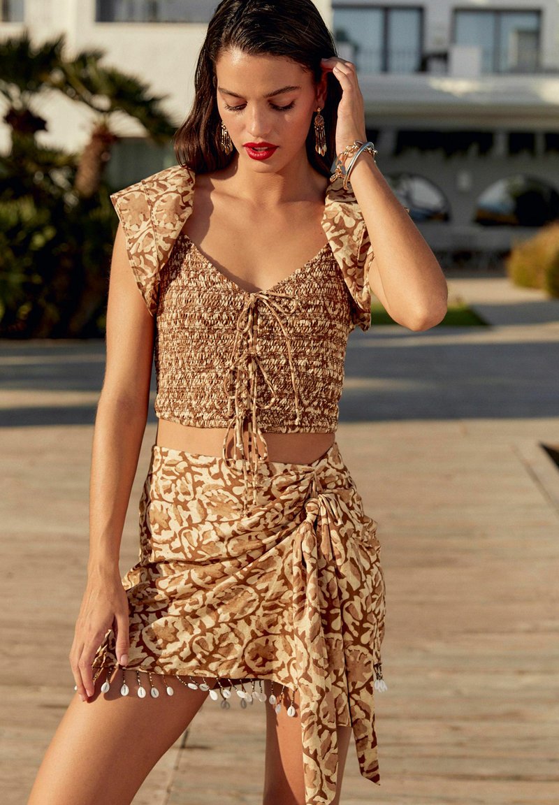 Bruine cropped top met bloemenpatroon, pofmouwen en trekkoordsluiting; bijpassende wikkelrok met parelaccenten aan de zoom.