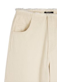 Beżowe bawełniane spodnie z postrzępionym dołem, boczną kieszenią i teksturowaną powierzchnią. Materiał wydaje się być trwały, oferując luźny krój.