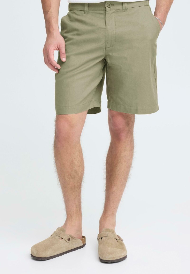 Homme portant un short vert olive avec des poches et des sabots en daim beige sans lacets, debout devant un fond uni.
