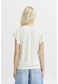 White short-sleeve top with ruffled accents on shoulders, soft fabric, relaxed fit, and round neckline, paired with denim shorts.