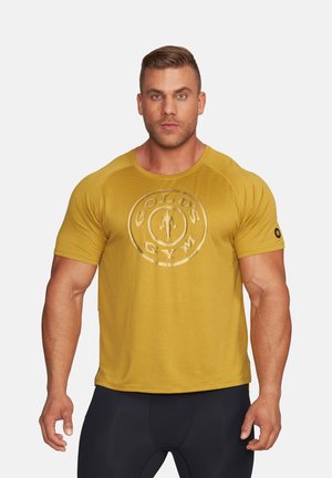 Muskularny mężczyzna zwrócony przodem, ubrany w musztardowożółtą koszulkę Gold's Gym i czarne spodnie treningowe na prostym tle.