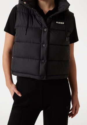 Veste sans manches matelassée noire avec capuche et boutons-pression, portée sur un top noir à manches courtes et un pantalon noir, main dans la poche.