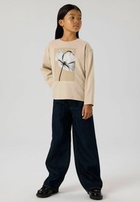 Beżowa bluzka z długim rękawem z czarno-białym wzorem kwiatowym; połączona z szerokimi ciemnoniebieskimi dżinsami i czarnymi butami.