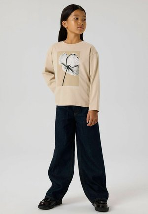 Beżowa bluzka z długim rękawem z czarno-białym wzorem kwiatowym; połączona z szerokimi ciemnoniebieskimi dżinsami i czarnymi butami.