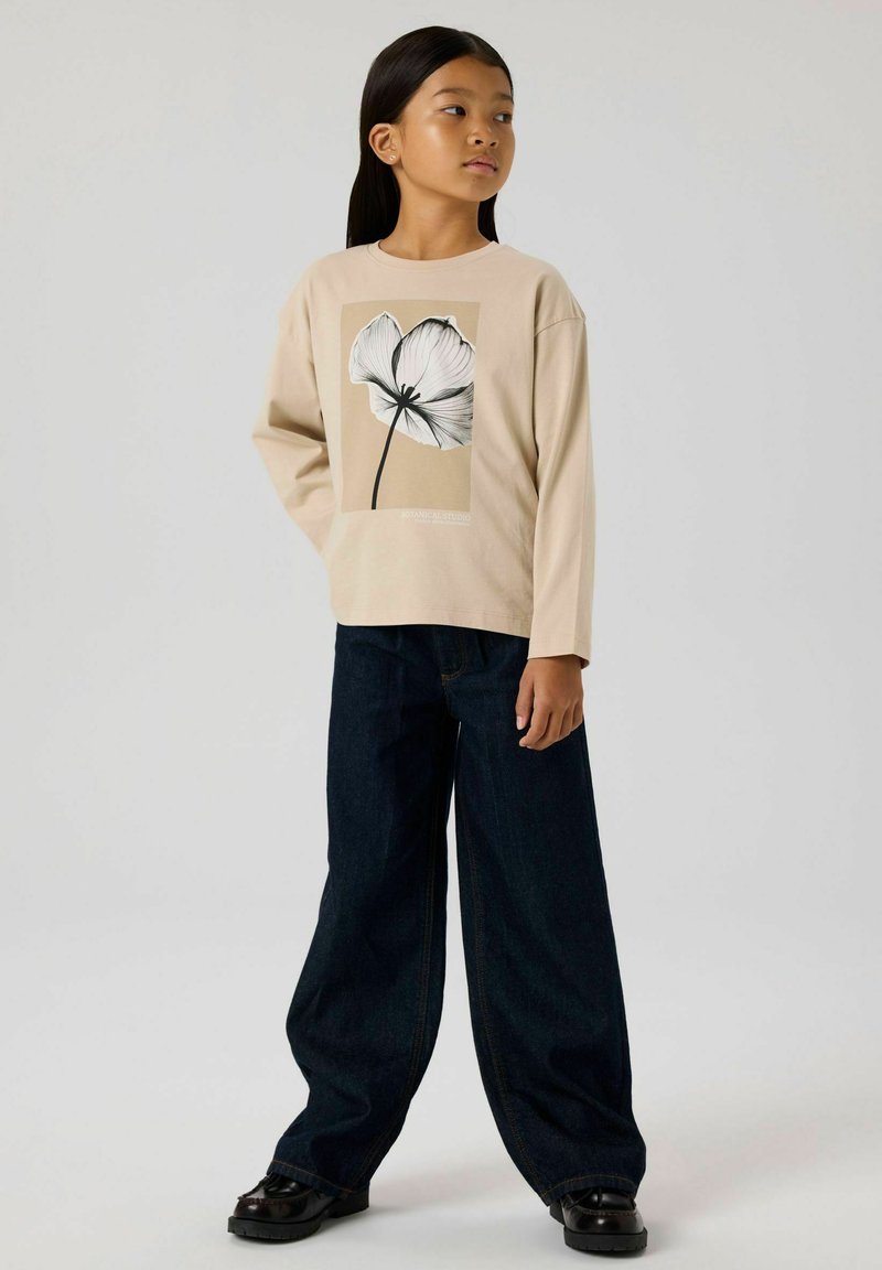 Beżowa bluzka z długim rękawem z czarno-białym wzorem kwiatowym; połączona z szerokimi ciemnoniebieskimi dżinsami i czarnymi butami.