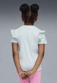 Niña con el cabello trenzado en dos moños, vestida con una camiseta de mangas acampanadas de color azul claro y pantalones rosas, de pie con las manos entrelazadas detrás de la espalda.