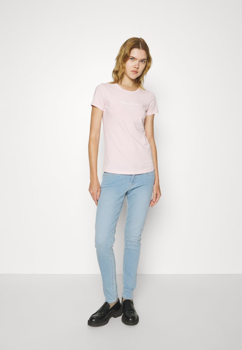 T-shirt ajusté rose clair à manches courtes et col rond, arborant un logo blanc imprimé sur la poitrine, associé à un jean slim bleu clair et des chaussures noires.