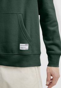 Sudadera verde oscuro con un bolsillo frontal, puños y dobladillo de canalé. Presenta una etiqueta blanca con texto en la parte inferior izquierda. Textura de tela suave.