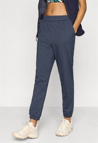 Σκούρες μπλε φόρμες με ελαστική μέση και μανσέτες, κατασκευασμένες από μαλακό υλικό. Συνδυασμένες με αθλητικά παπούτσια σε ουδέτερους τόνους για σπορ εμφάνιση.