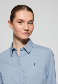 Jasnoniebieska koszula zapinana na guziki z klasycznym kołnierzykiem, z małą granatową haftowaną figurką na lewej piersi. Gładki bawełniany materiał.