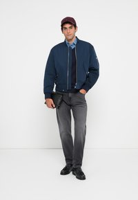 Man wearing a navy bomber jacket, blue shirt, gray jeans, black tassel loafers, burgundy cap, holding a black clutch bag, standing against white background.