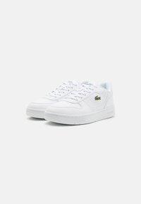 Hvite lær sneakers med teksturert overflate, rund tå, snøring foran, og grønt Lacoste-logo på siden.