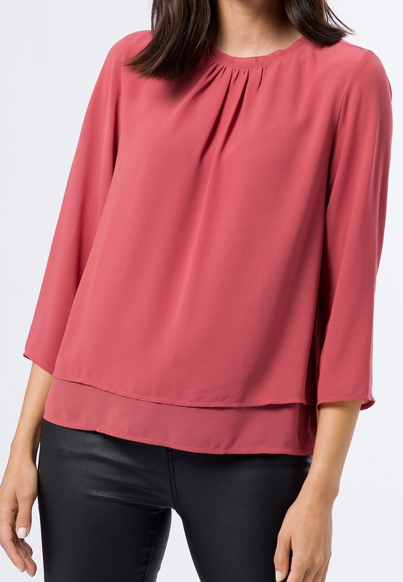Femme portant un chemisier ample à manches longues couleur rose poussiéreuse avec des plis au niveau de l'encolure et un pantalon noir ajusté sur un fond clair.