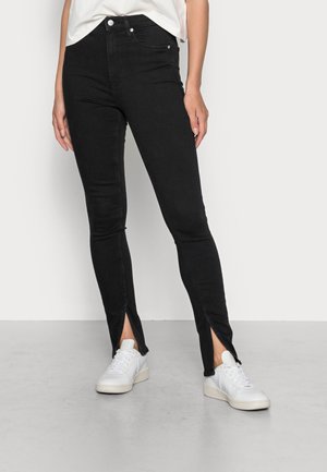 Person iført sorte high-waisted skinny jeans med frontankelåbninger og hvide sneakers, stående mod en ensartet lys baggrund.