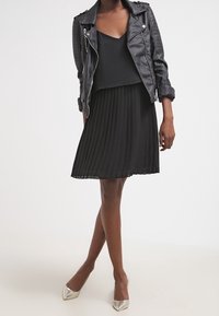 Jupe plissée noire associée à un top sans manches et une veste en cuir noire de motard. La tenue est complétée par des talons argentés brillants.