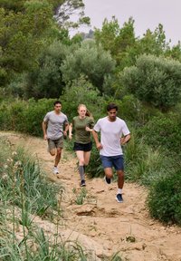 Kolme juoksijaa hiekkapolulla: yksi harmaassa paidassa ja shortseissa, toinen vihreässä paidassa ja mustissa shortseissa, ja kolmas valkoisessa paidassa ja sinisissä shortseissa.