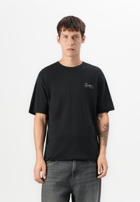 Ung mann iført en svart "Dystopia" t-skjorte og mørke jeans, stående mot en enkel hvit bakgrunn med en tatovering på venstre underarm.