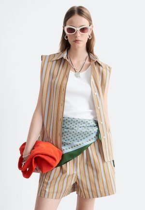 Femme portant un haut sans manches rayé et un short, débardeur blanc, ceinture en foulard à motif, lunettes de soleil blanches, bijoux argentés, tenant un sac orange vif.