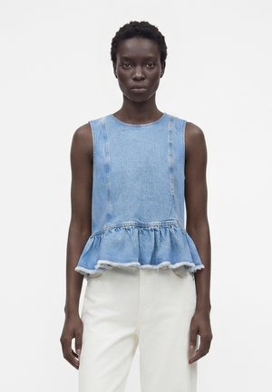 Femme à la peau foncée portant un haut peplum en denim bleu clair sans manches et un pantalon blanc taille haute, posant devant un fond blanc uni.