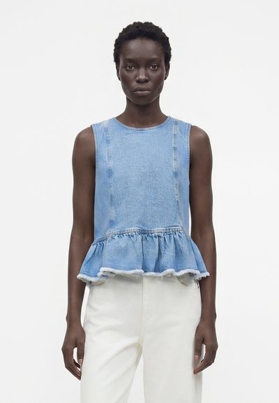 Femme à la peau foncée portant un haut peplum en denim bleu clair sans manches et un pantalon blanc taille haute, posant devant un fond blanc uni.