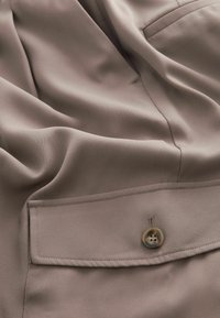 Veste en tissu marron clair avec un rabat de poche à boutons, des coutures apparentes et de doux plis dans le matériau.