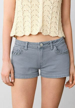 Femme portant un short en jean bleu clair avec des poches avant lacées et un haut crème en crochet sans manches, debout les mains détendues.