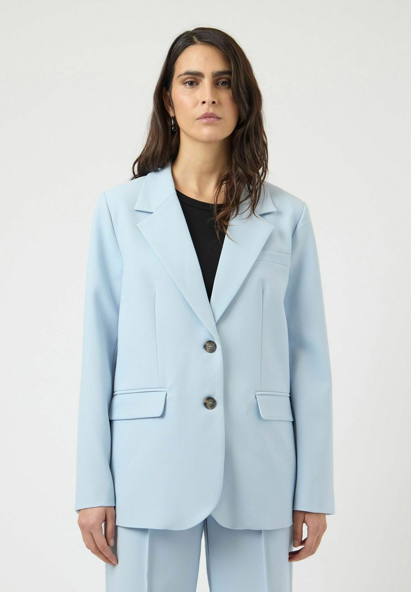 Γυναίκα με μακριά σκούρα μαλλιά, φορούν ανοιχτό μπλε σακάκι προσαρμοσμένης γραμμής και ταιριαστό παντελόνι πάνω από μαύρη μπλούζα, στέκεται μπροστά σε λευκό φόντο.