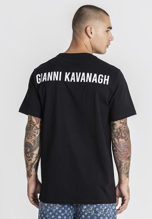 Uomo con tatuaggi su entrambe le braccia che indossa una maglietta nera con la scritta "GIANNI KAVANAGH" in lettere bianche sulla parte superiore della schiena.