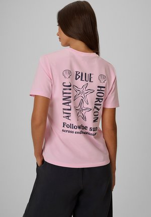 Kobieta z długimi ciemnymi włosami nosi jasno różowy t-shirt z rozgwiazdami, muszelkami i tekstem o Atlantyku, niebieskim horyzoncie i słońcu na plecach.