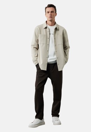 Lichtbeige overhemd met knopen en twee borstzakken, gedragen over een wit T-shirt, gecombineerd met donkerbruine broeken en witte sneakers.