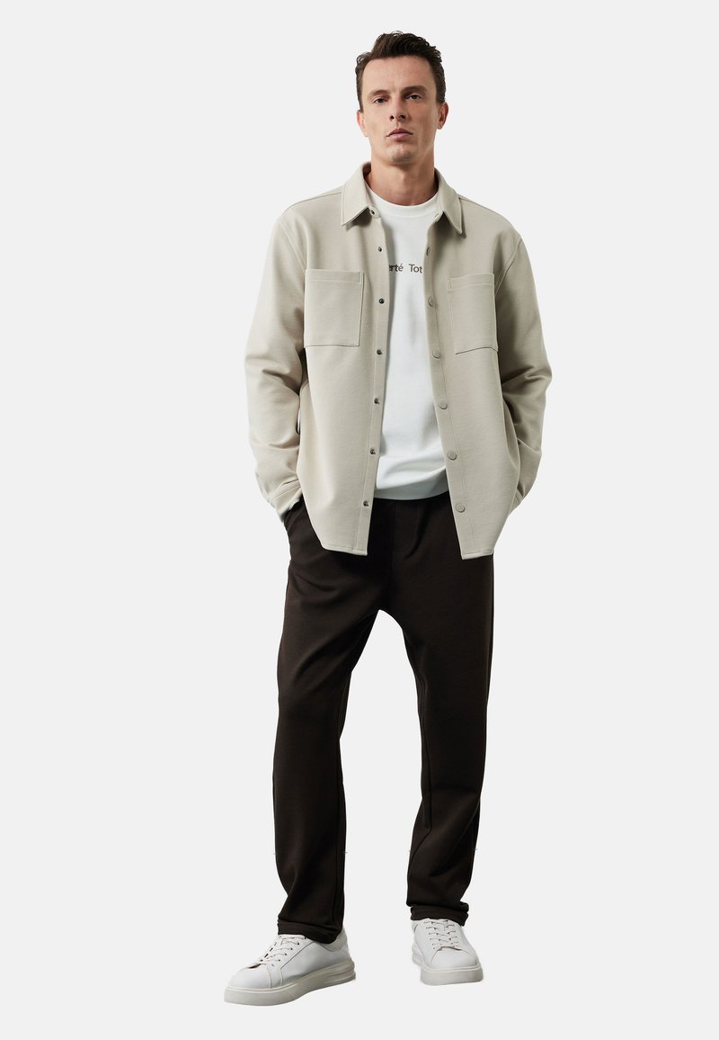 Lysbeige skjorte med knapper og to brystlommer, lagdelt over en hvid t-shirt, parret med mørkebrune bukser og hvide sneakers.